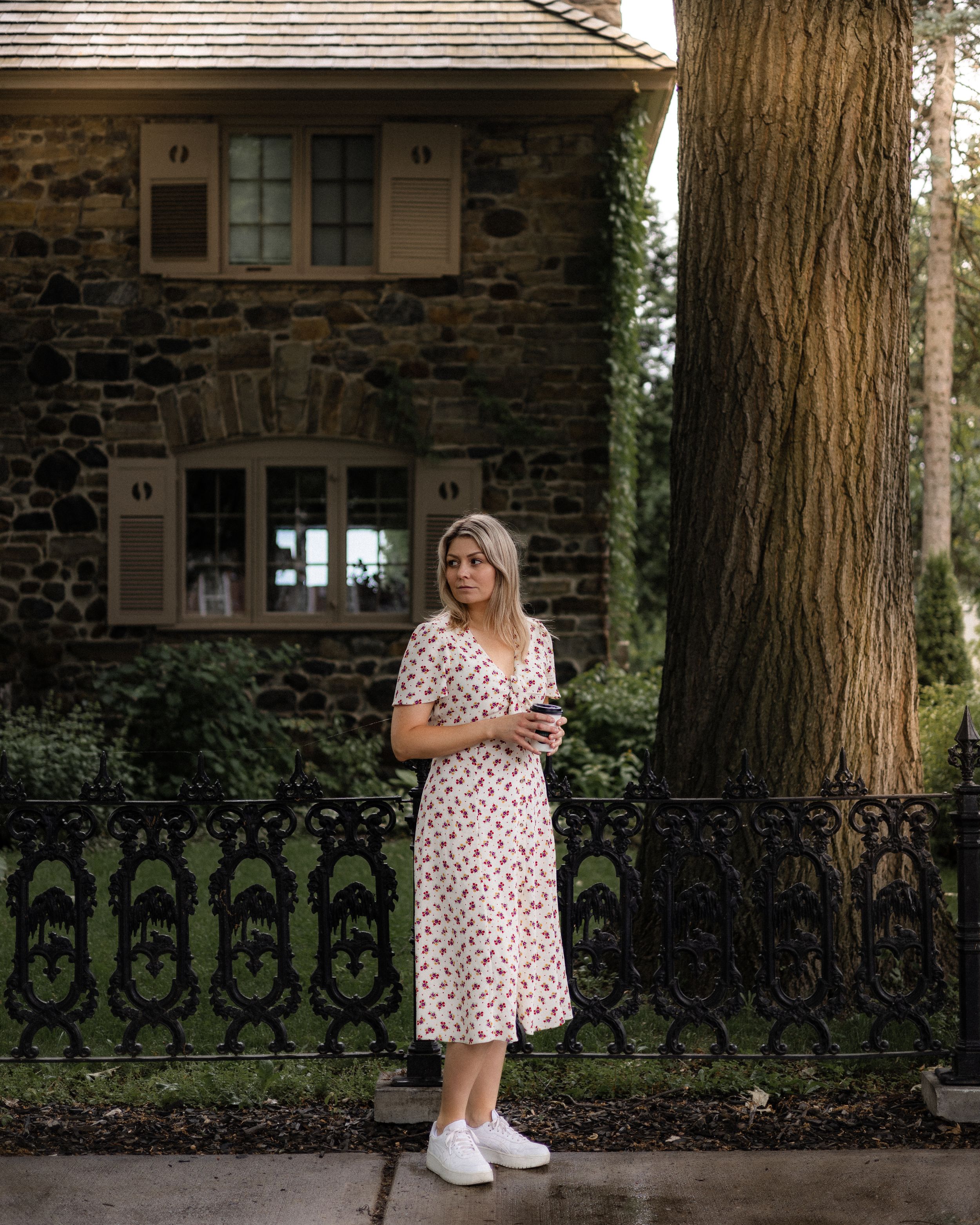 Woman in polka dot dress stands near stone house with iron fence.
