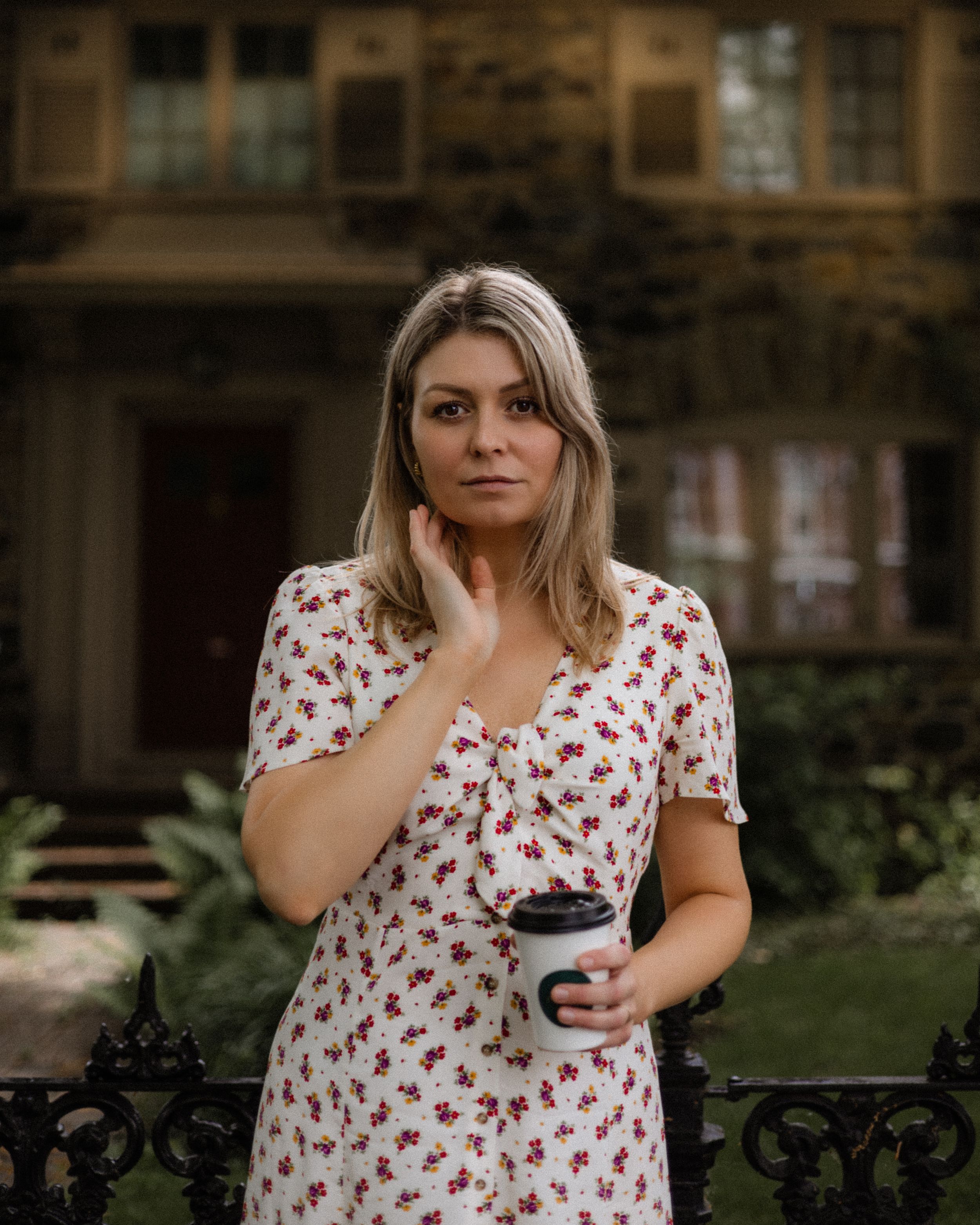 Woman in floral dress holding coffee cup poses outside historic building.