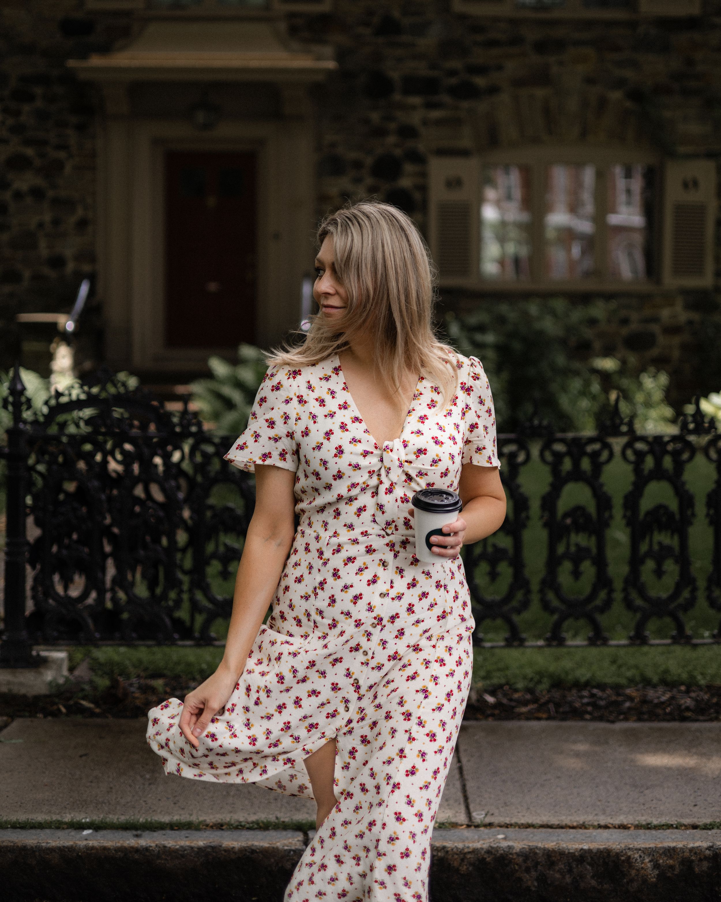 Woman in floral dress poses by ornate iron fence outside historic stone building.