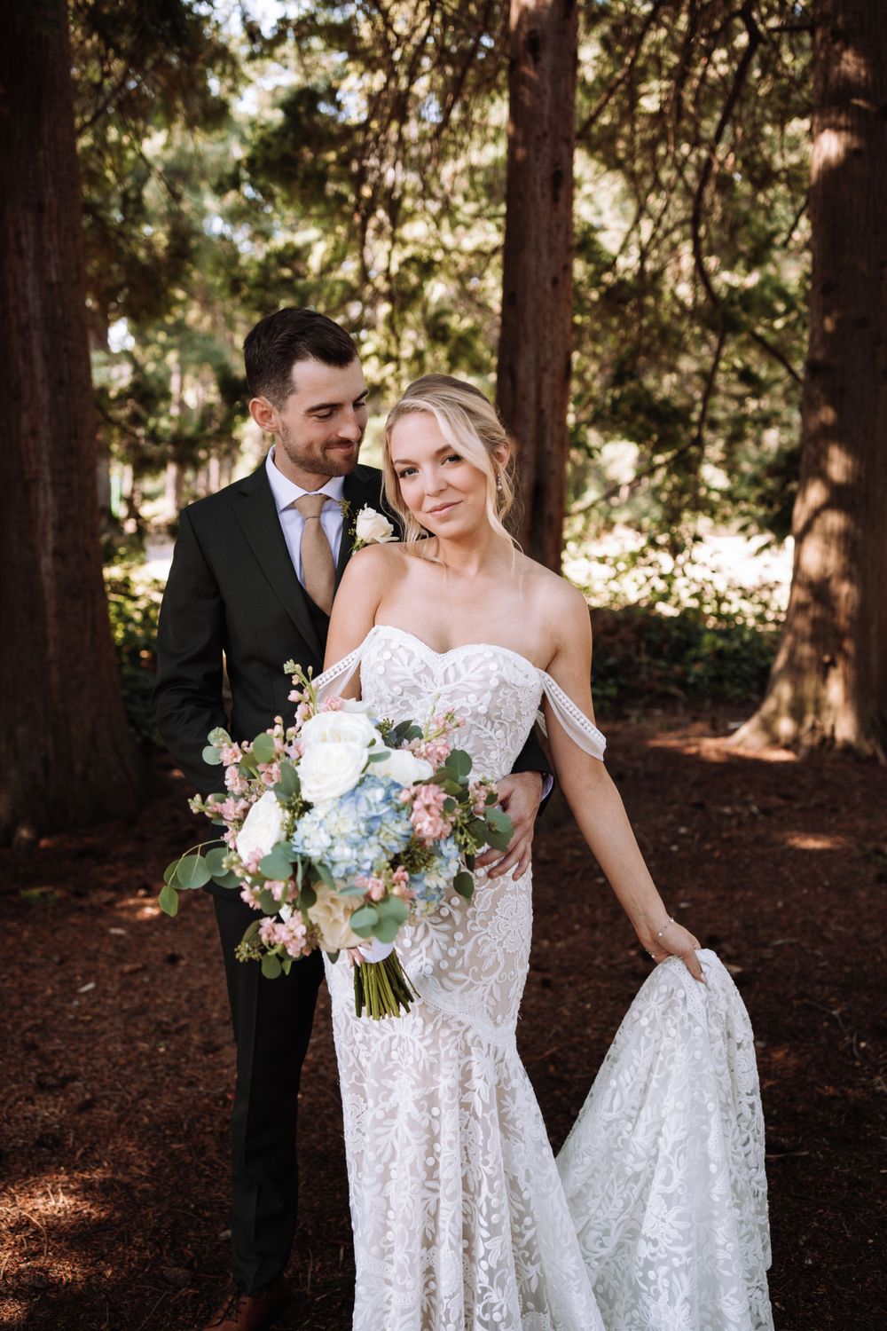 A couple poses in a forest setting wearing wedding attire with a beautiful bouquet of mixed flowers.