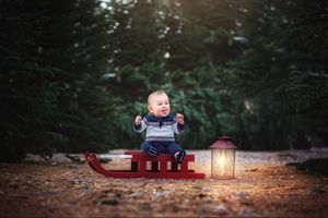 A child bundled in winter clothing sits on a vintage red sled next to a glowing lantern at dusk.