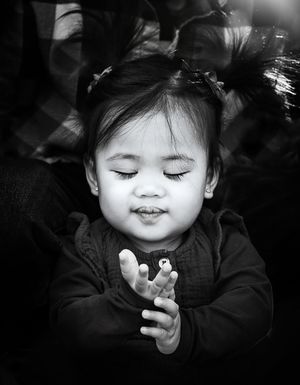Black and white portrait of a smiling toddler with closed eyes and clasped hands.