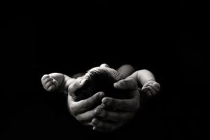 A dramatic black and white image of hands cradling a newborn baby against dark background.