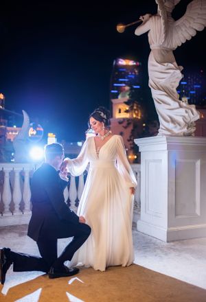 A romantic proposal scene at night with an illuminated angel statue and city lights in the background.