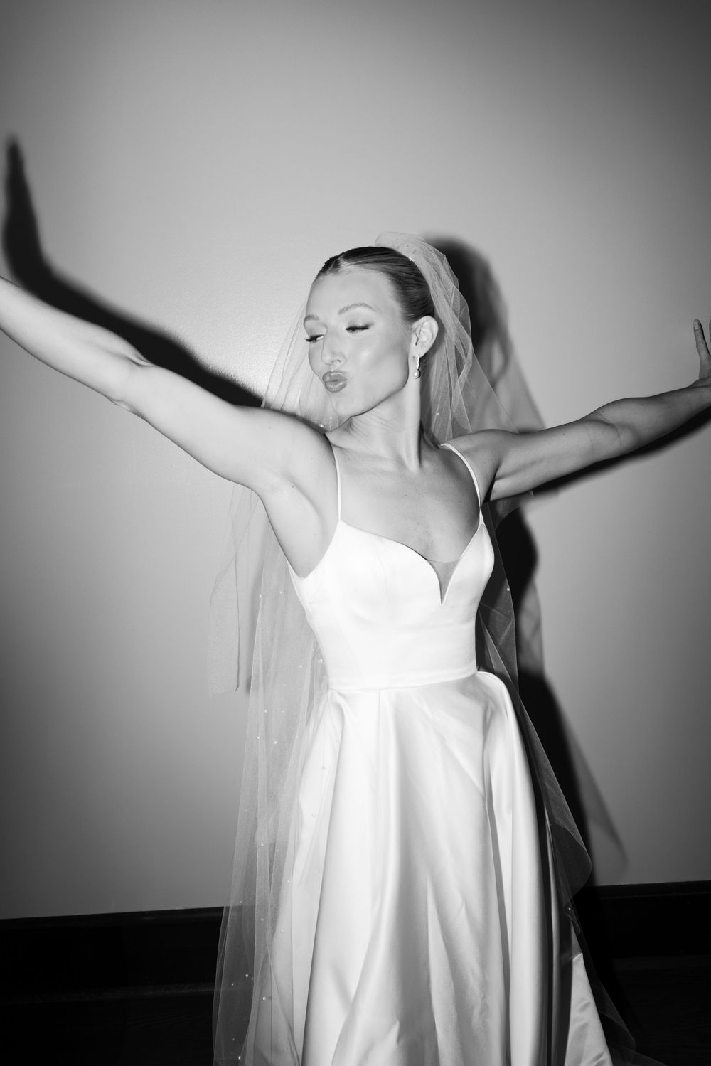 A person in a white wedding dress with a veil strikes a dramatic pose with arms spread wide against a black and white backdrop.