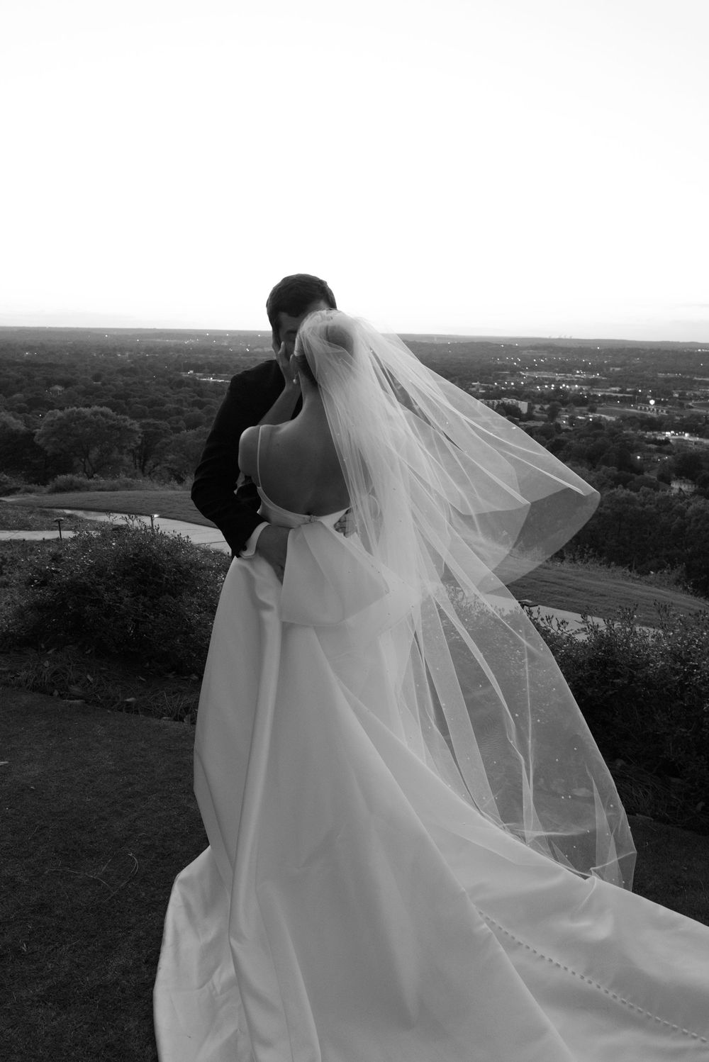 Bride's flowing veil catches wind as couple embraces on hilltop overlooking scenic landscape at dusk.
