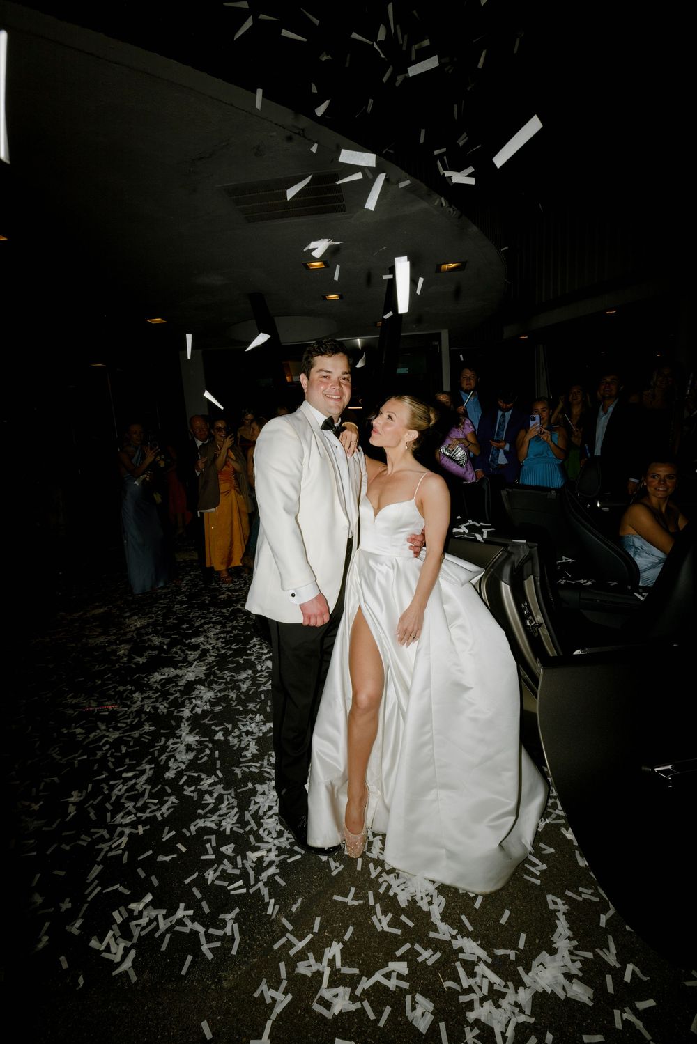 Two people in white formal attire celebrate at their wedding reception with confetti falling around them on a dark dance floor.