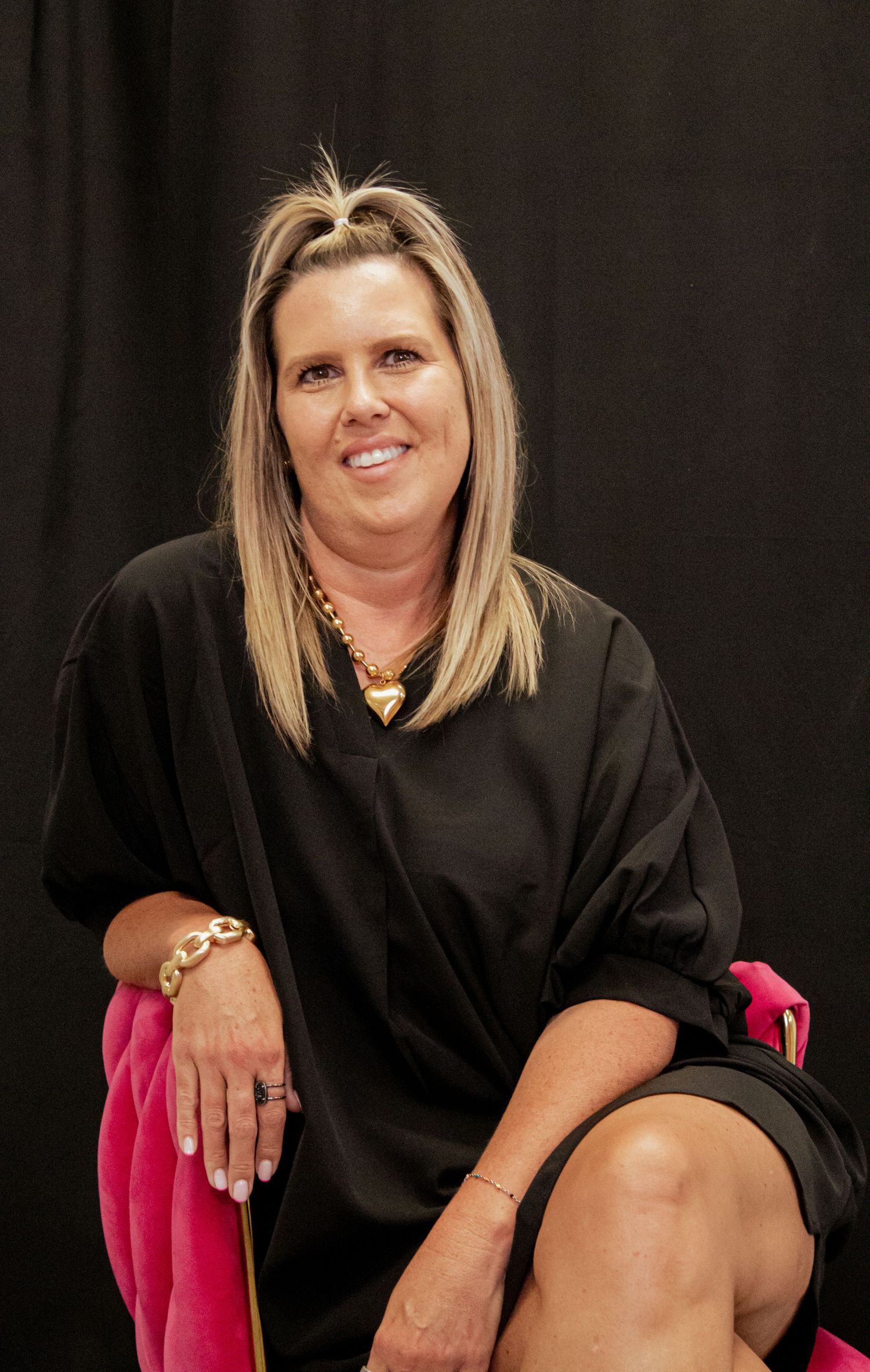 Professional portrait of a person with blonde hair wearing a black top while seated in a pink chair against a dark background.