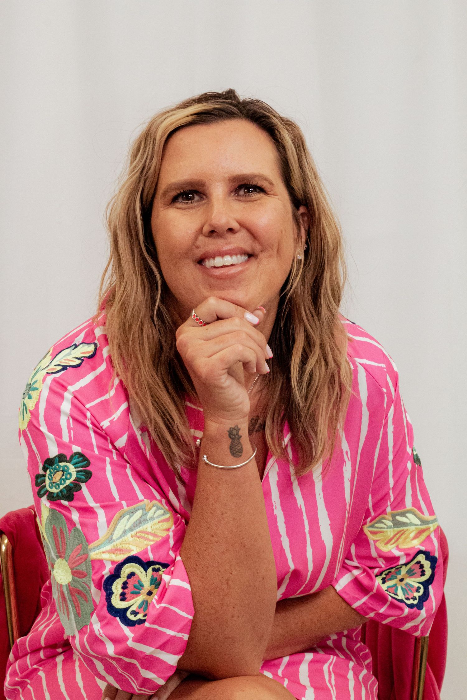 A smiling person in a vibrant pink floral robe with blonde hair poses with hand on chin against a white background.