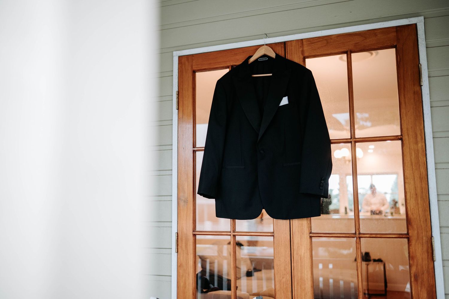 A black blazer jacket hangs on a wooden door frame against a white wall in natural lighting.