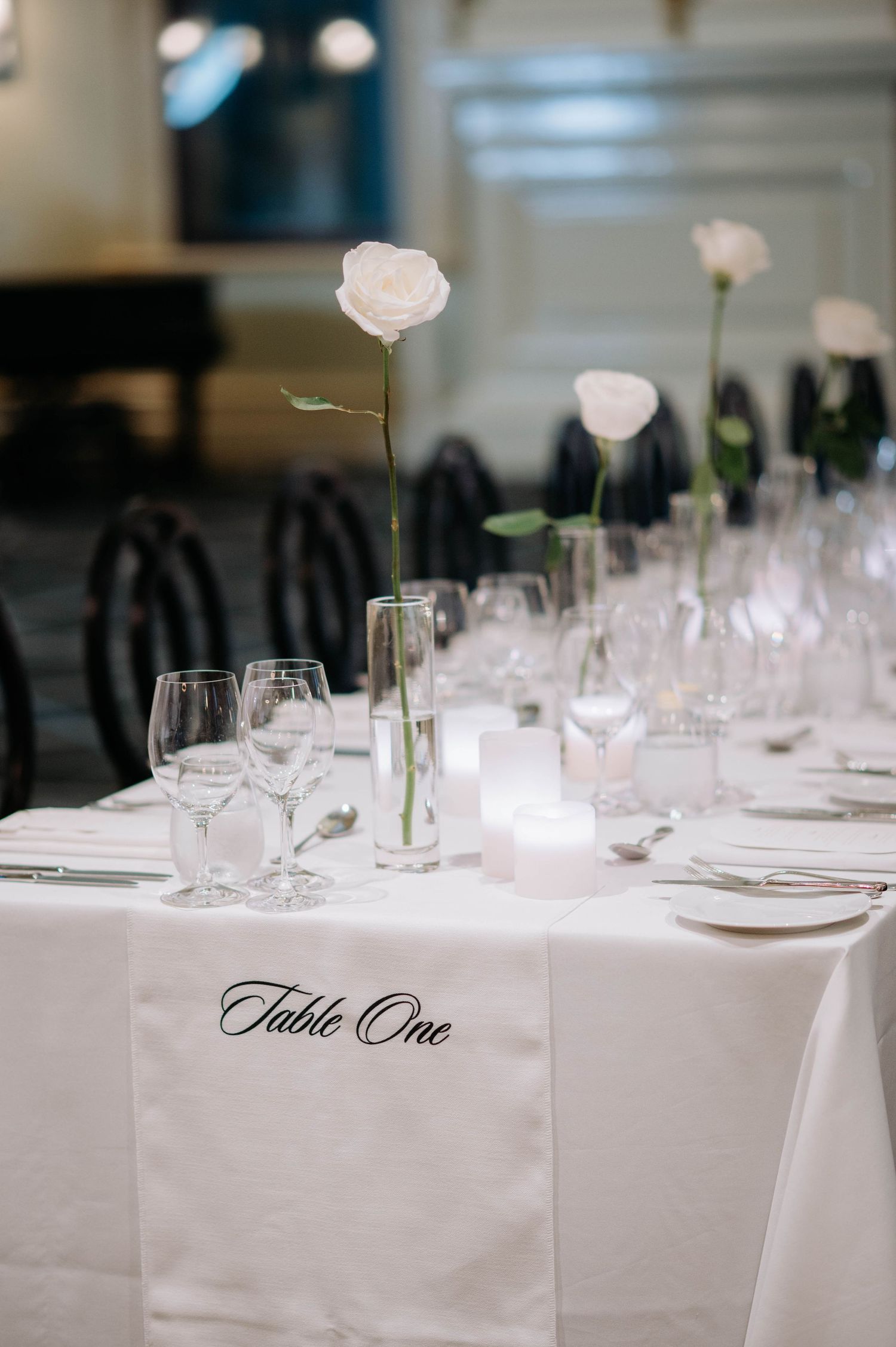 Elegant wedding table setting with crisp white linens, wine glasses, and single white roses in minimalist vases.