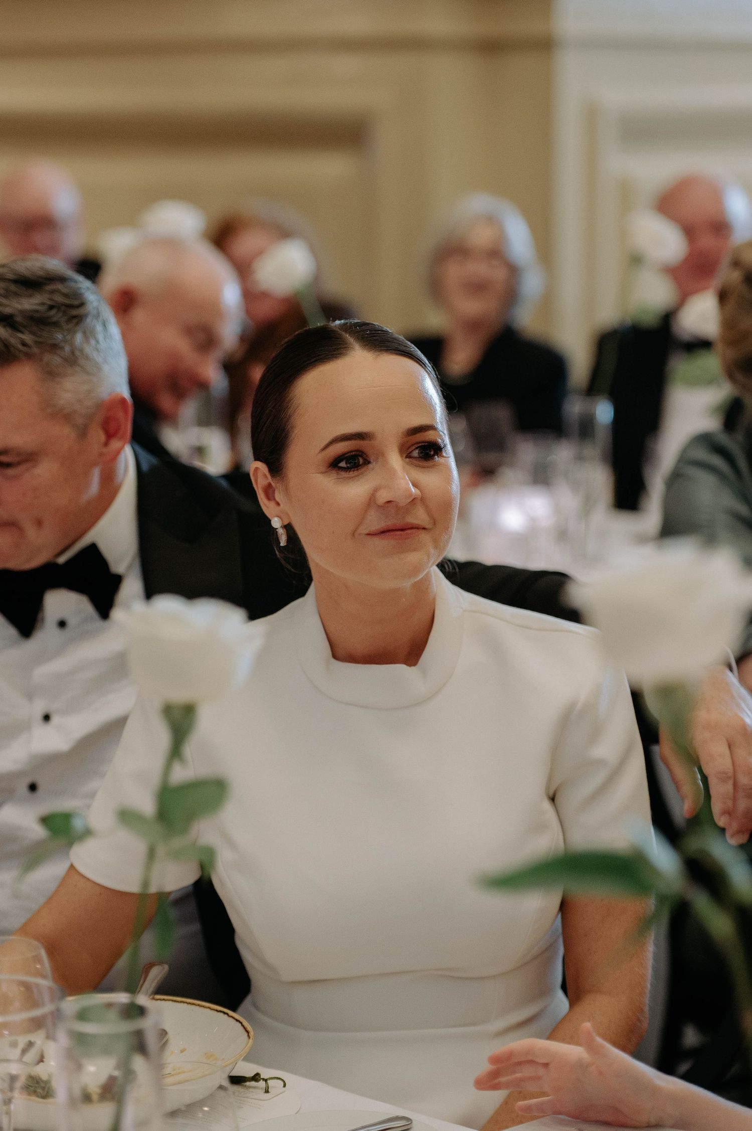 Elegant woman in white dress smiles while seated at formal dinner event with other guests in evening attire.
