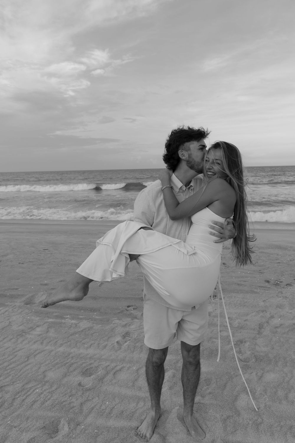 Black and white photo of groom lifting and kissing bride on a sandy beach at sunset.