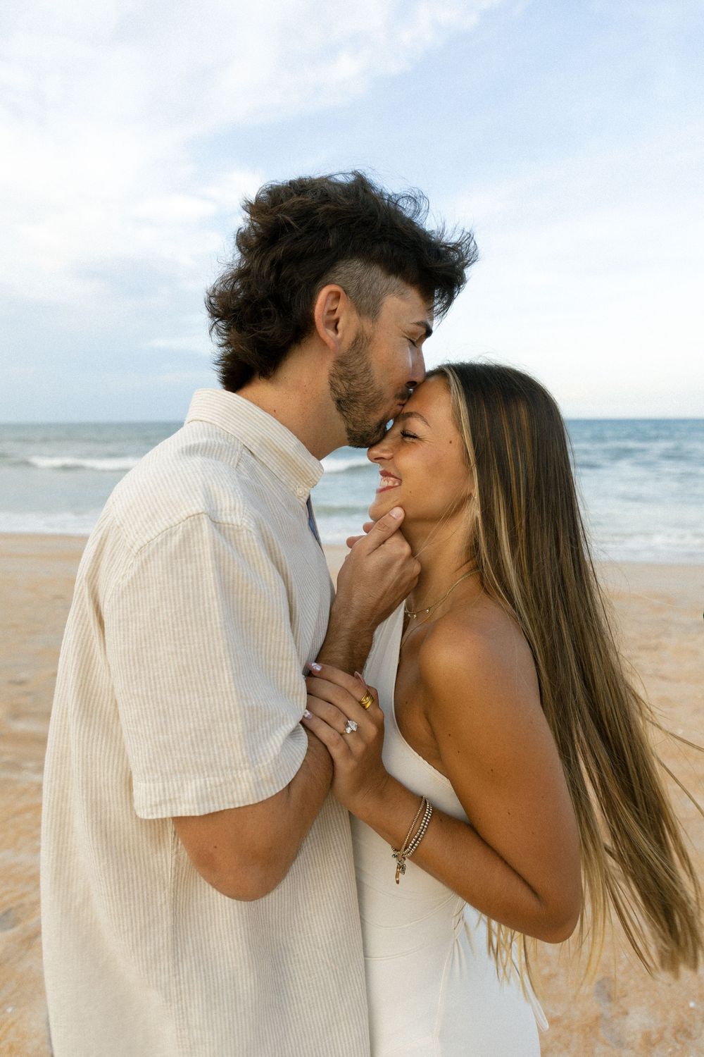 Romantic couple almost kissing on sandy beach at sunset, woman wearing white dress and engagement ring.