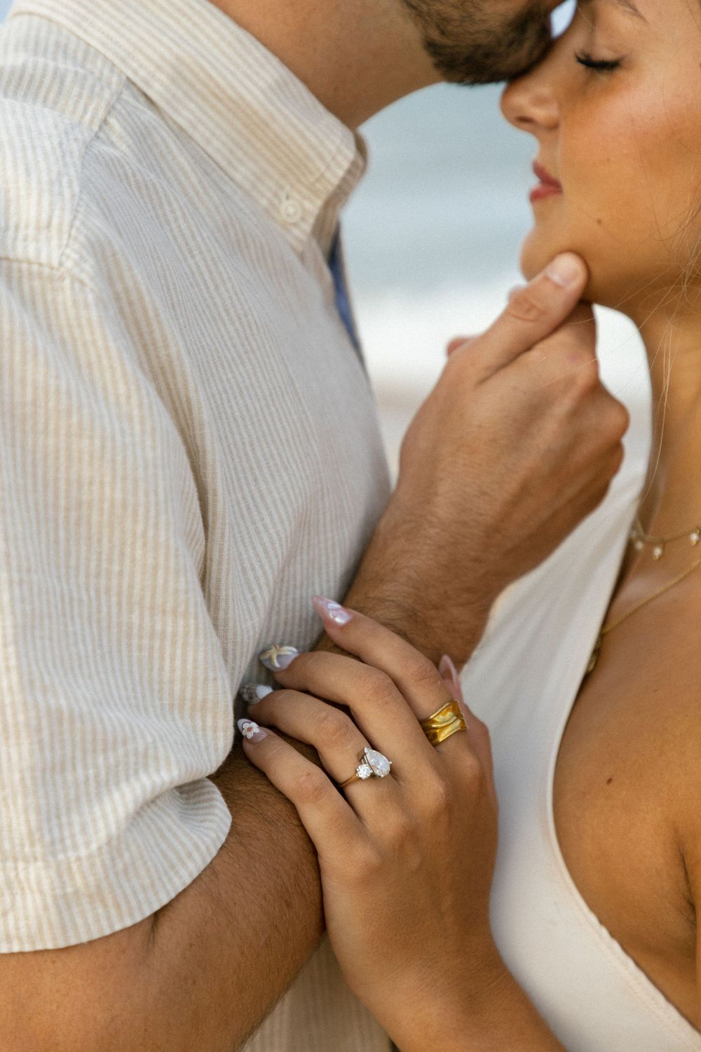 Close-up of engaged couple almost kissing at beach, woman showing diamond engagement ring and gold band.