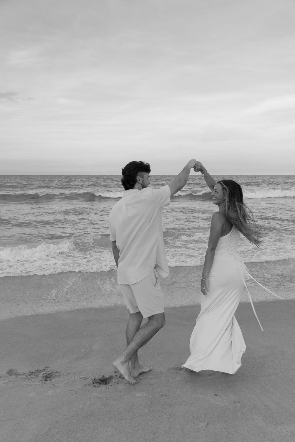 Black and white photo of couple dancing on beach at sunset, man twirling woman in white dress.