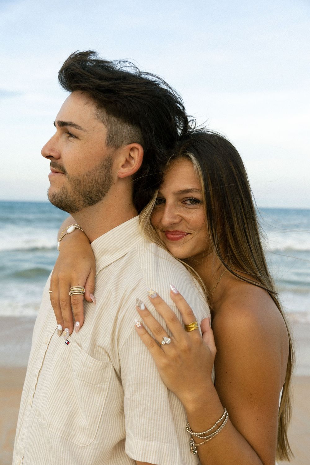 Engaged couple embraces on the beach at dusk, woman showing off diamond engagement ring and gold jewelry.