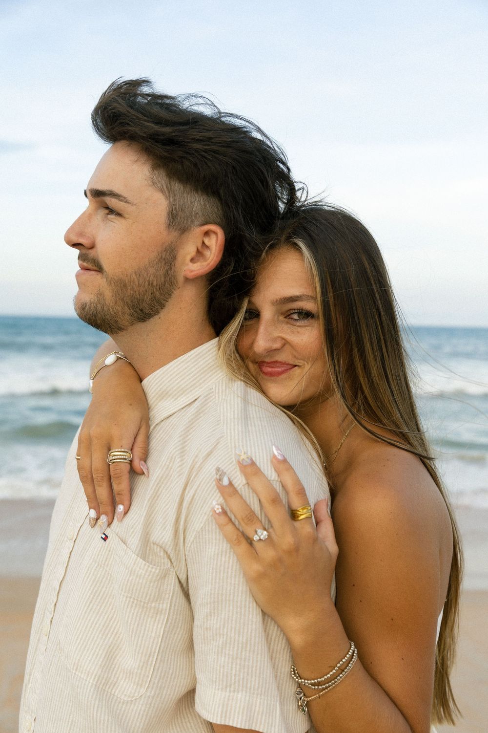 Couple embracing on beach, woman showing engagement ring, ocean waves in background.