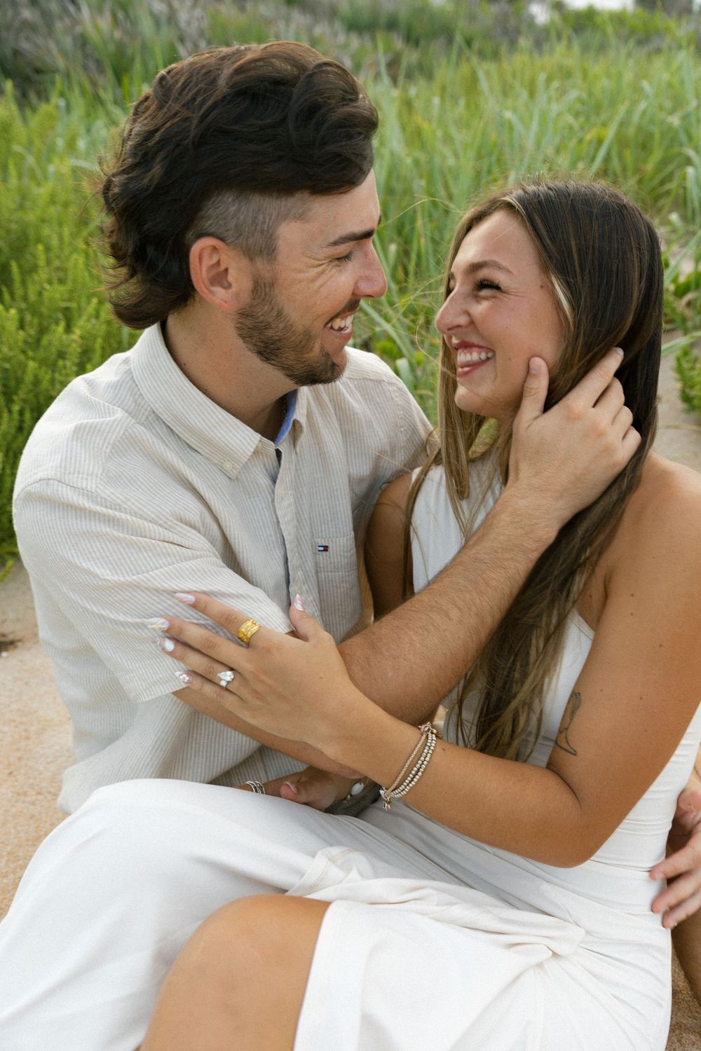 Happy couple laughing and gazing at each other while sitting outdoors near green foliage.