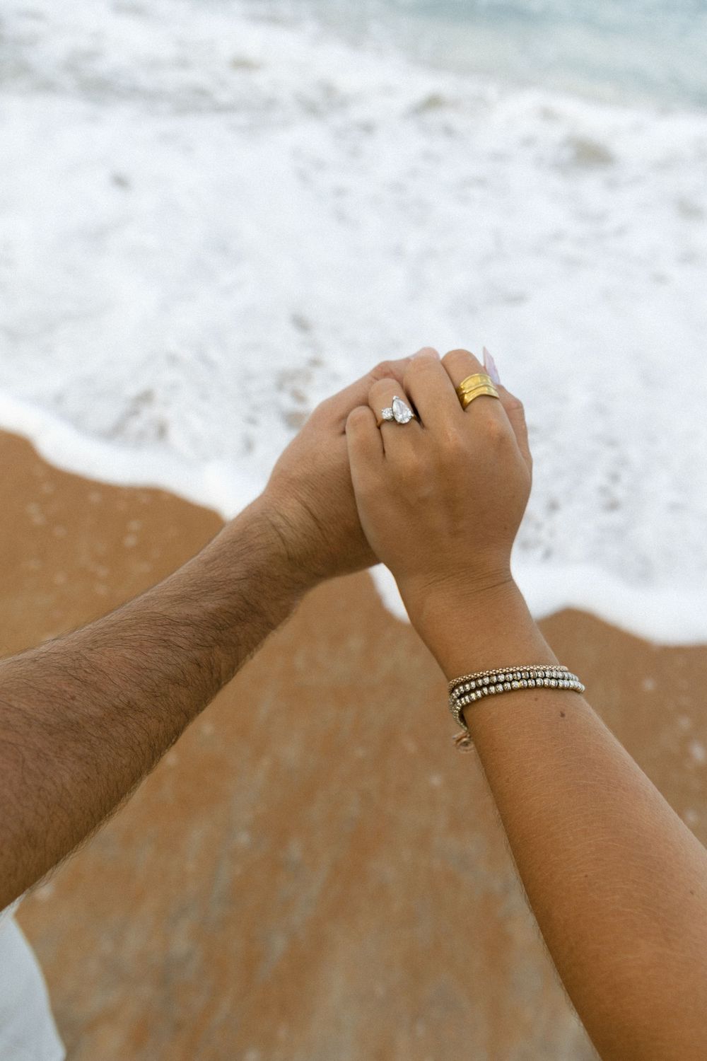 Engaged couple holding hands on beach shoreline, woman wearing diamond engagement ring and gold band.