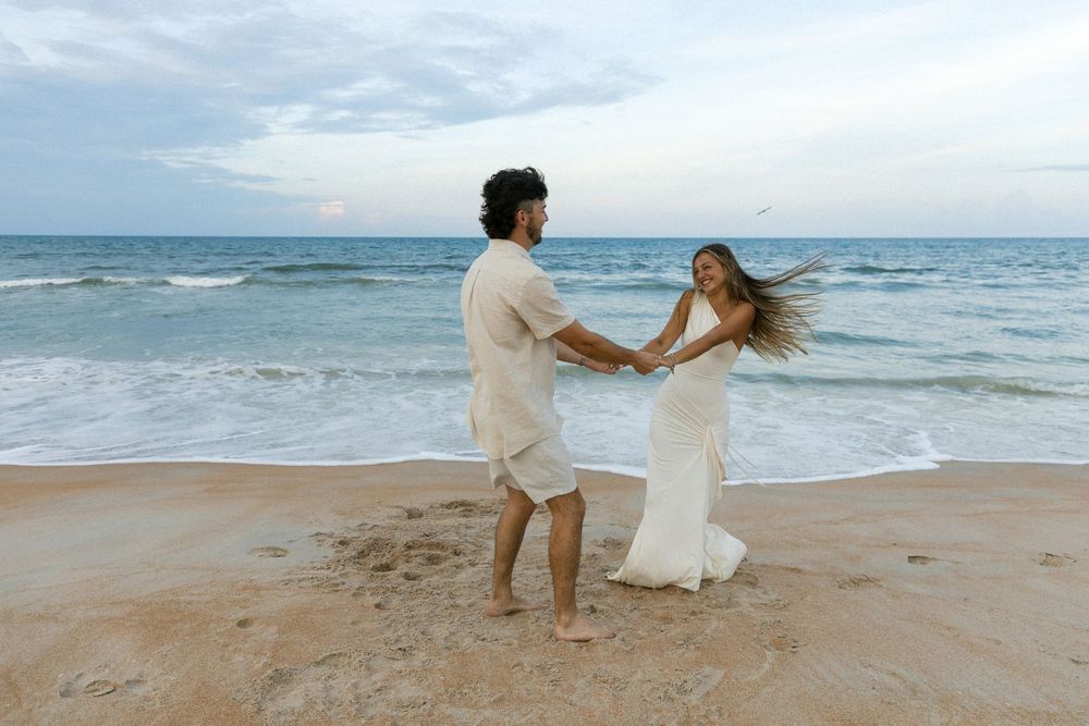 Joyful couple dancing and holding hands on sandy beach with ocean waves behind them.