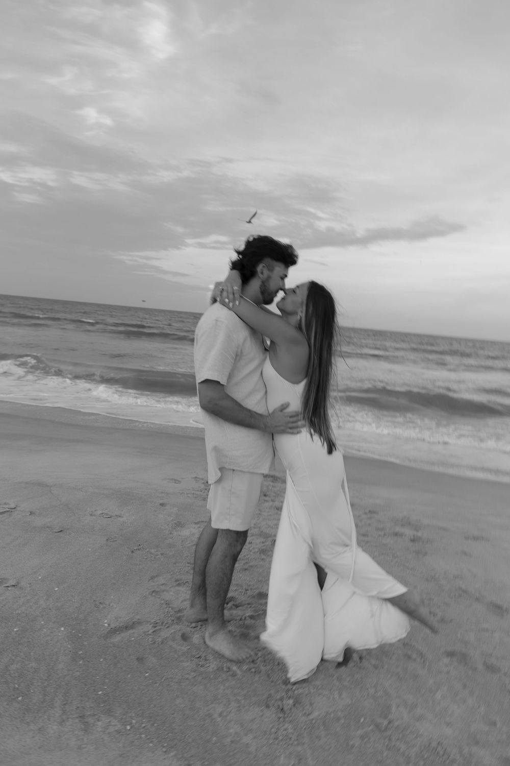 Black and white photo of couple kissing on beach at sunset, woman in white dress.