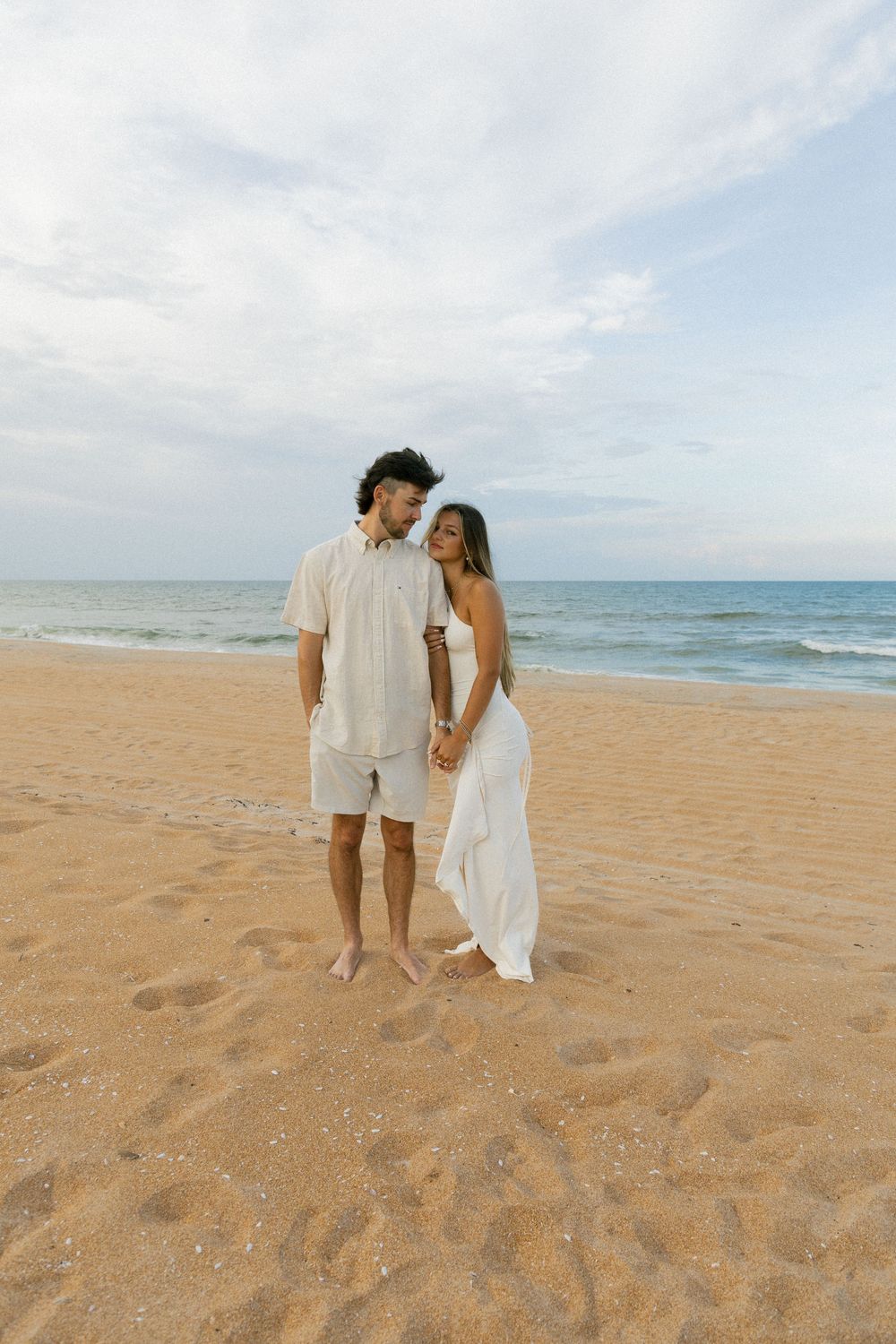 Happy engaged couple walking hand in hand on sandy beach, woman in flowing white dress.