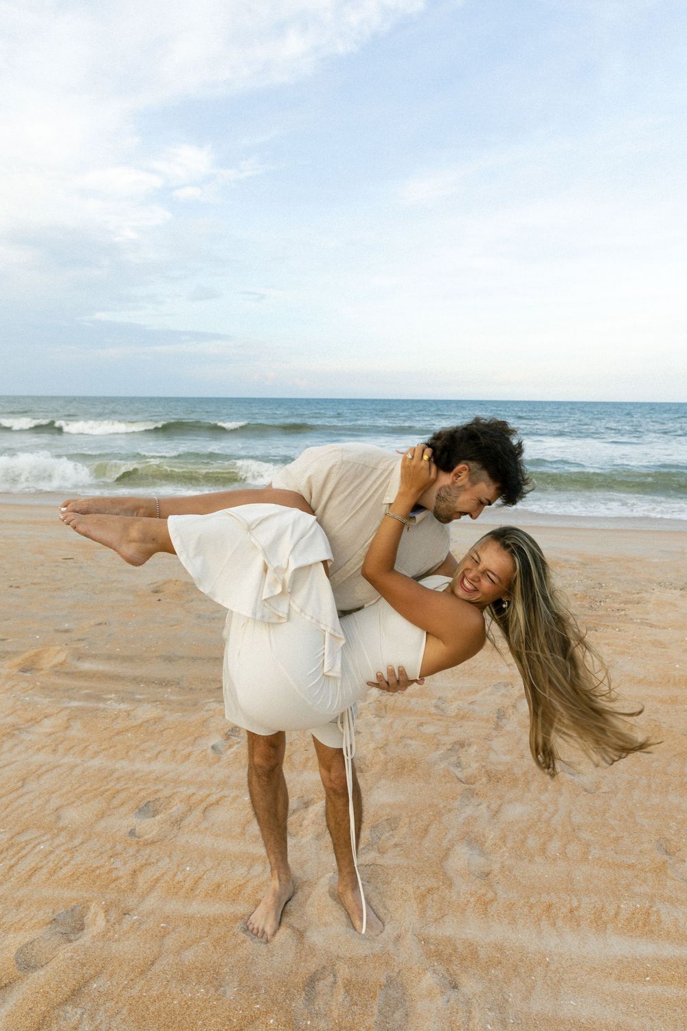 Man dipping and kissing woman on sandy beach, her hair blowing in the breeze.
