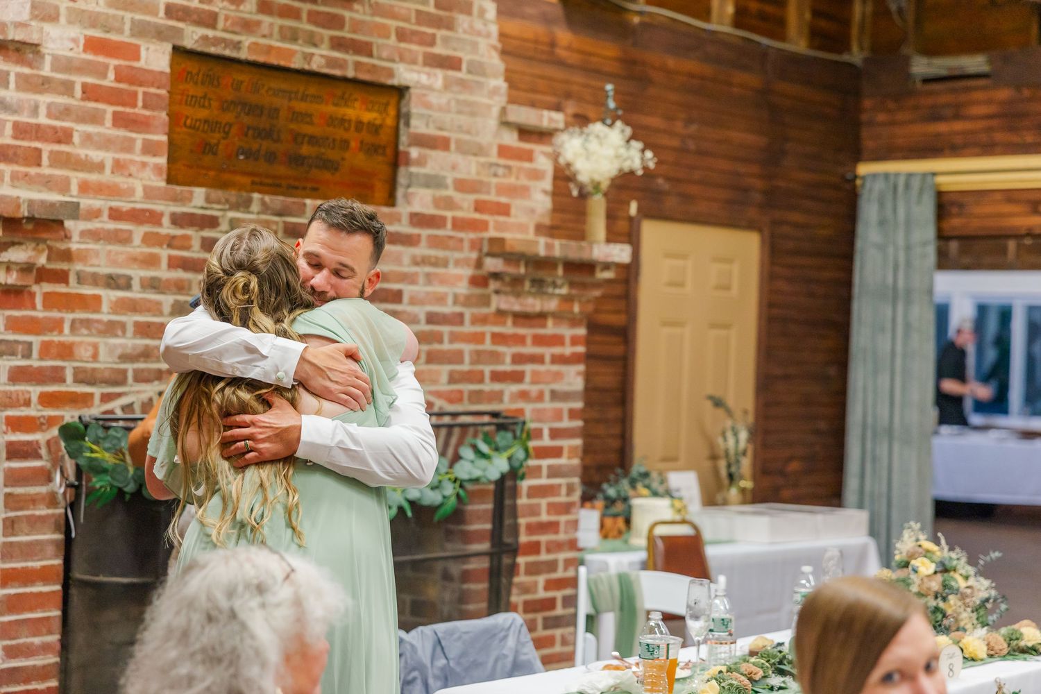 Wedding party dances in a rustic venue with exposed brick walls and strung lights overhead during reception celebration.