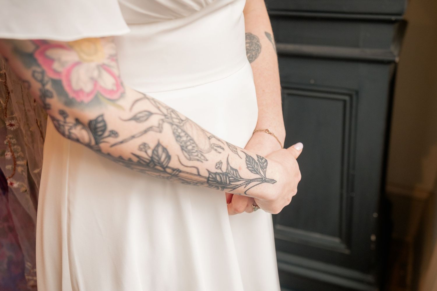 A tattooed arm cradles a pregnant belly draped in white fabric against a dark wall.