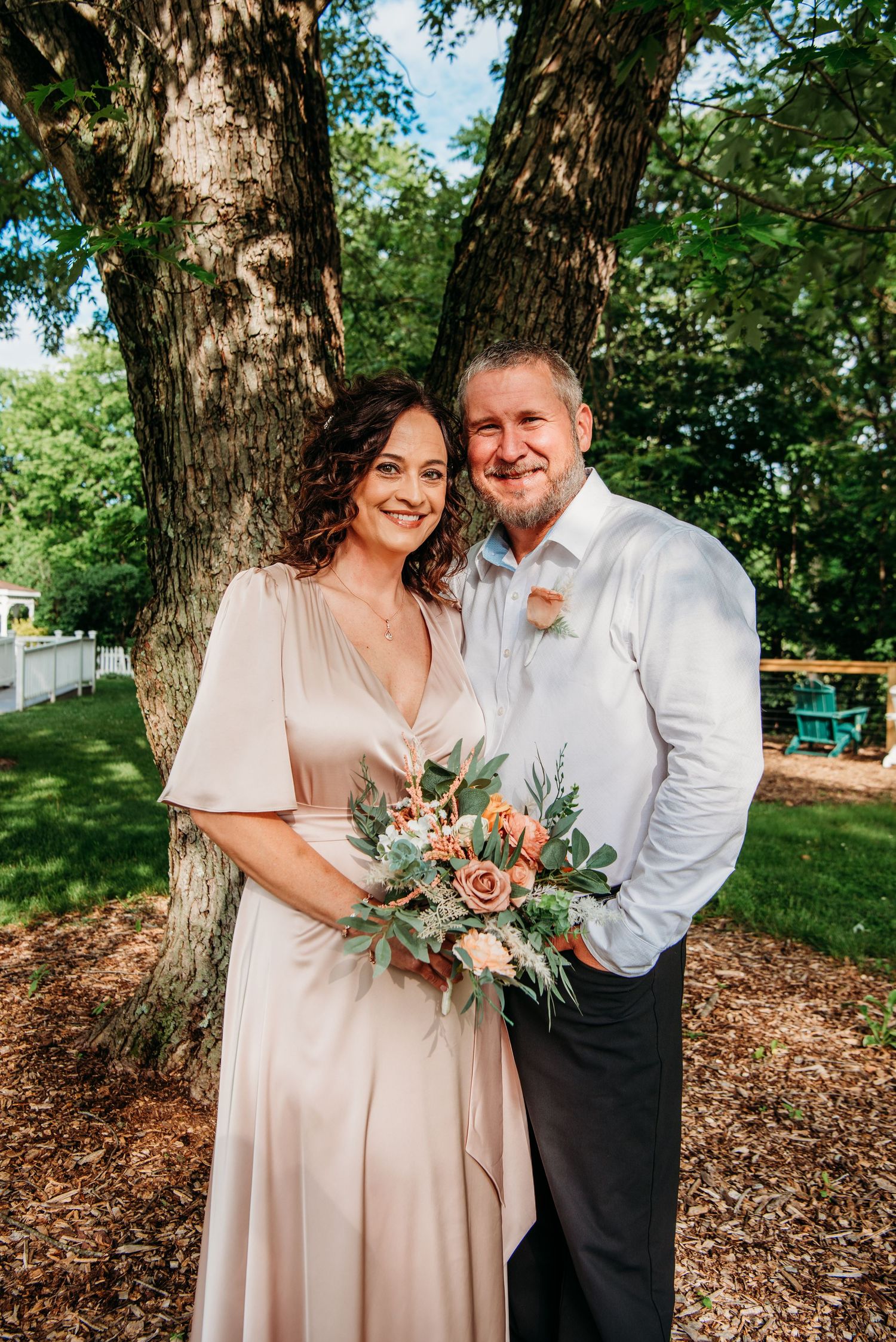 A couple poses outdoors holding a peach and blush floral bouquet with greenery at the Appalachian Wedding Chapel.