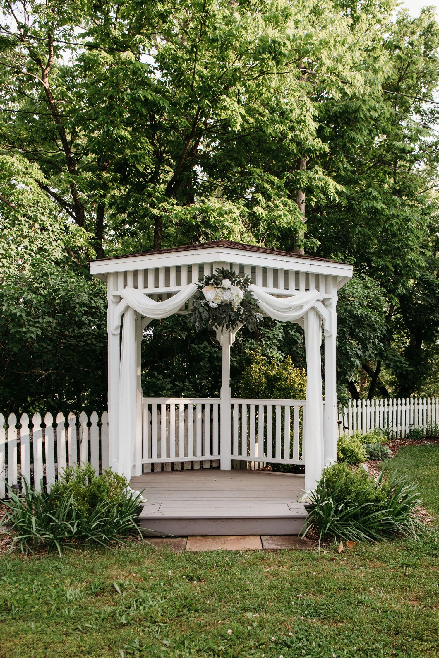 A white garden gazebo decorated with flowing fabric and floral arrangements stands among lush greenery at the Appalachian Wedding Chapel.