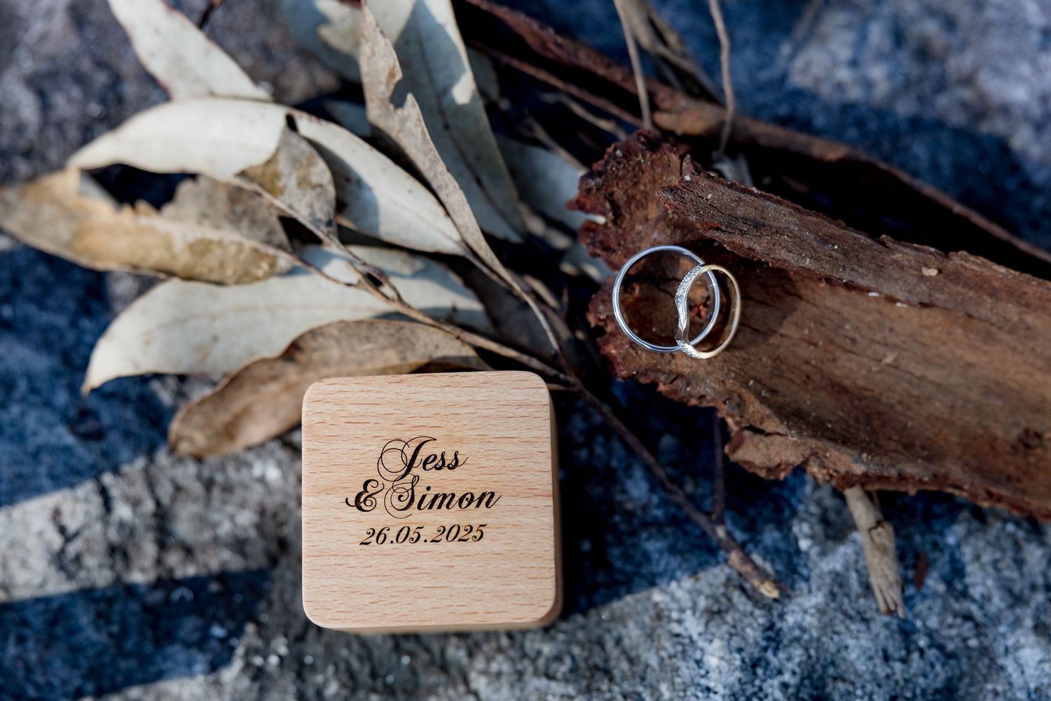 Wedding rings displayed on rustic wood with names engraved on wooden box and dried leaves in romantic setting.