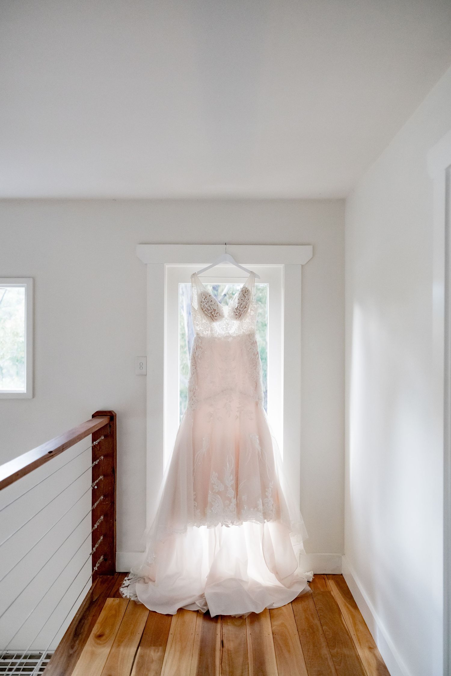 A blush pink wedding dress with lace details hangs elegantly in a white hallway near a staircase with hardwood floors.