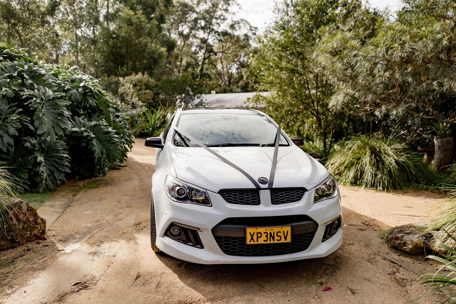 A white Holden HSV sports car parked on a dirt path surrounded by tropical plants and trees.