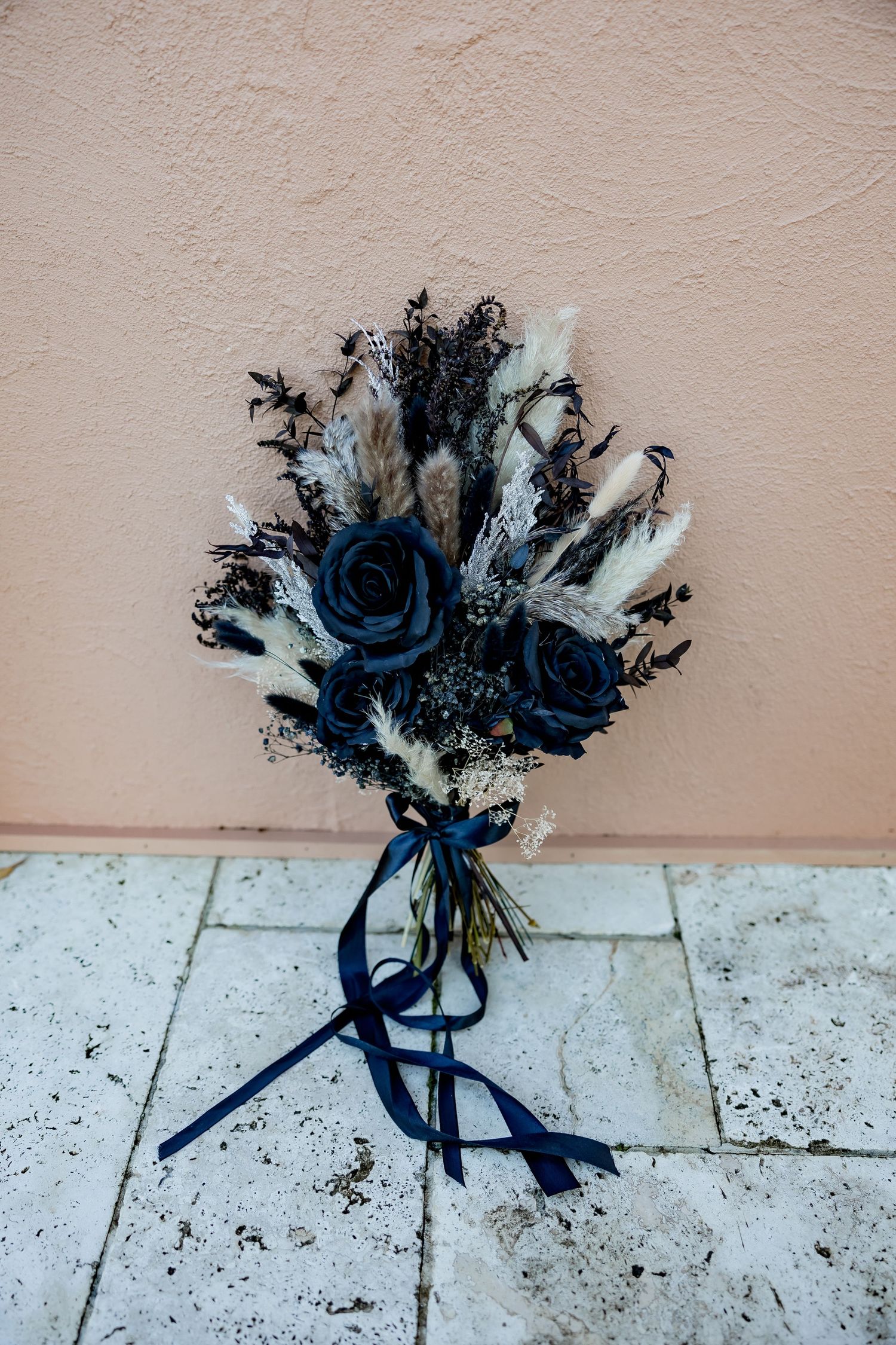 A moody wedding bouquet featuring dark blue roses and feathers tied with navy ribbon against a textured wall.