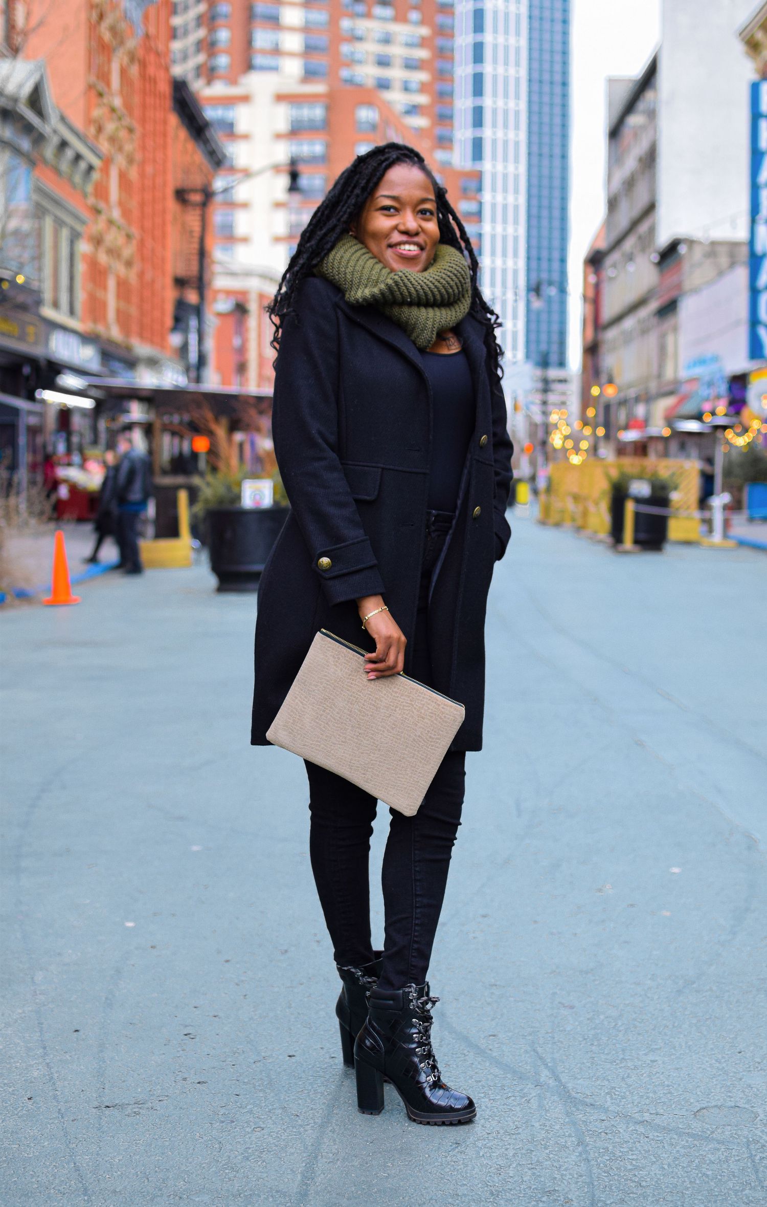 Stylish person in black winter coat with green scarf carrying clutch on city street with tall buildings in background.