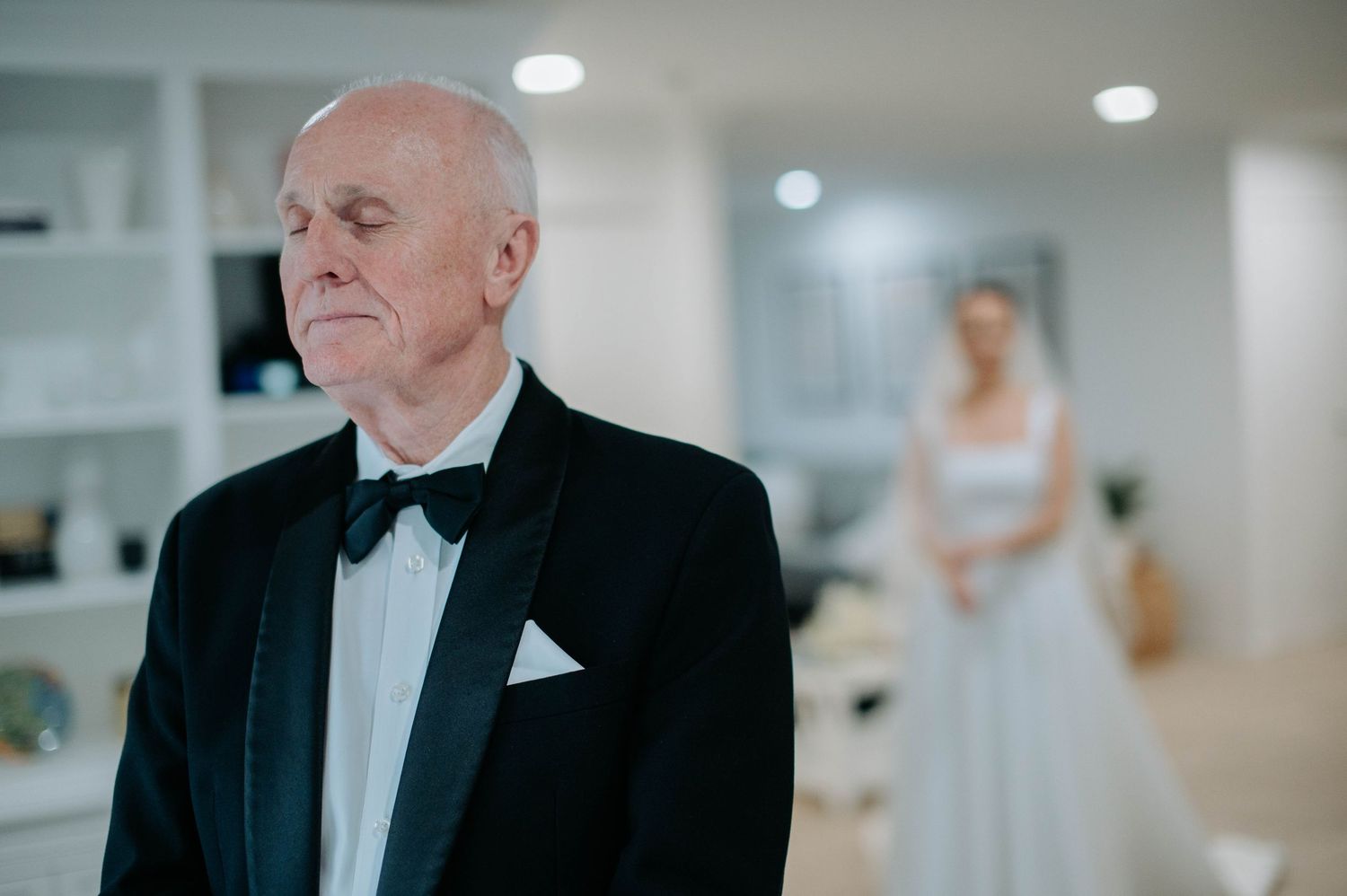 Elegant older man in black tuxedo and bowtie stands in bright room with someone in white dress visible in background.