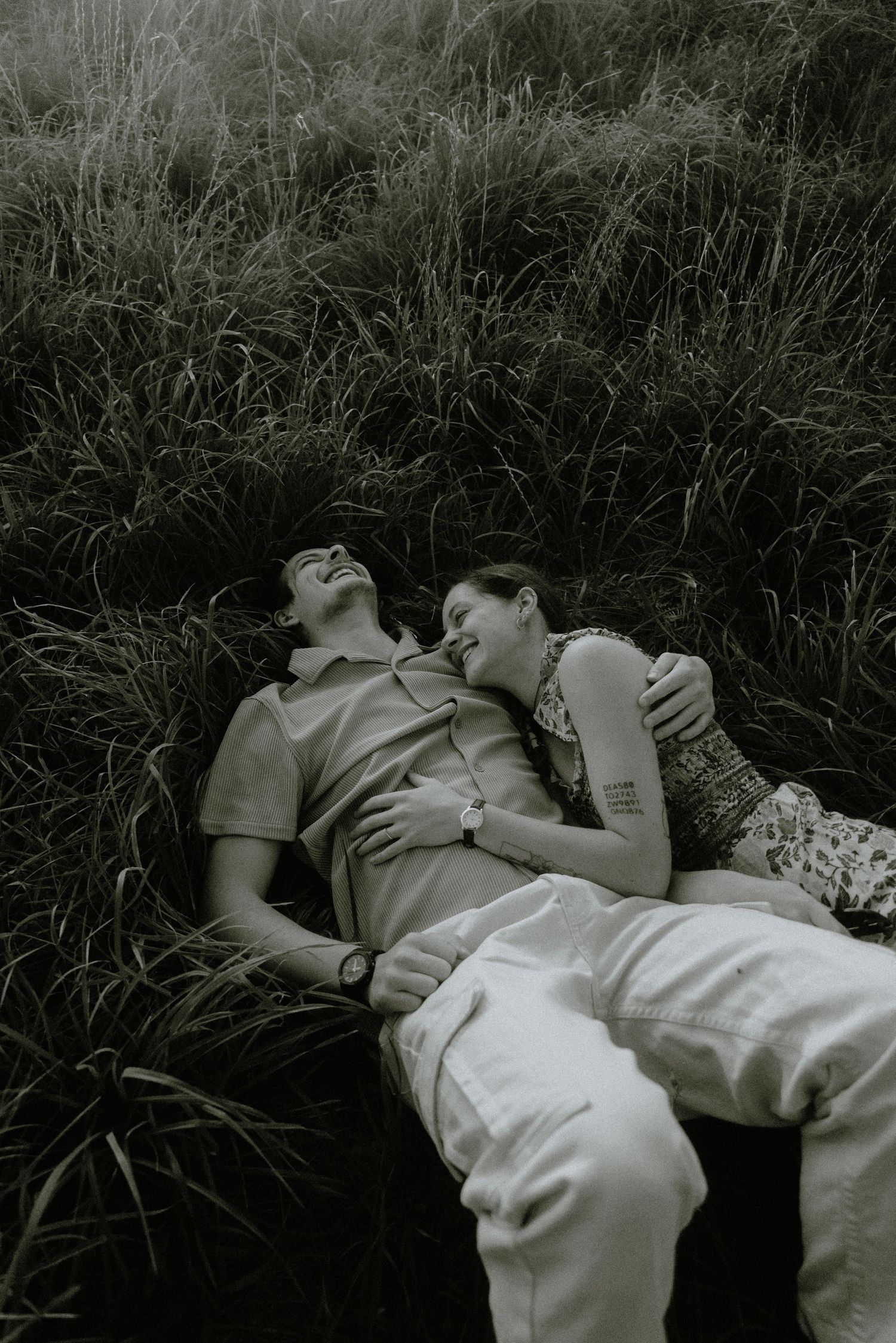 A couple rests intimately together in tall grass, captured in black and white photography.