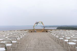 Wedding ceremony setup with white chairs and floral arch on a stone terrace overlooking the ocean.