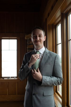 Someone in a light gray suit adjusting their tie near wooden-framed windows.