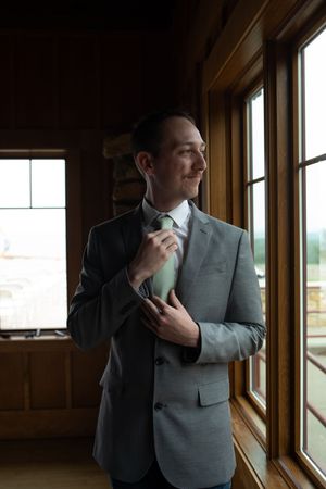A figure in formal attire standing by large windows in warm lighting.