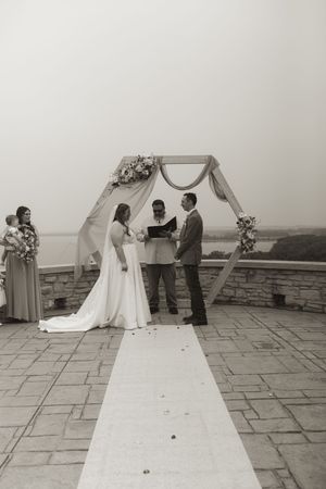 An outdoor wedding ceremony setup with a hexagonal arch and white runner on a stone patio overlooking mountains.