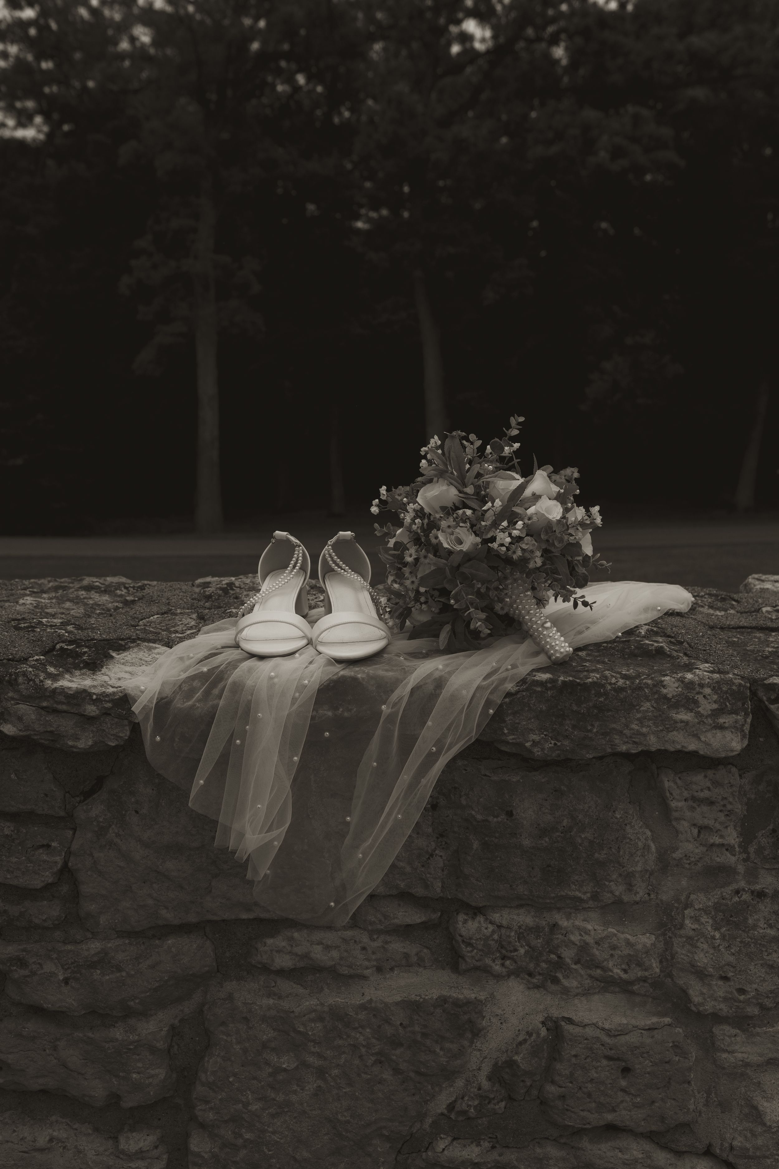 Black and white image of bridal accessories and bouquet arranged on stone wall outdoors.