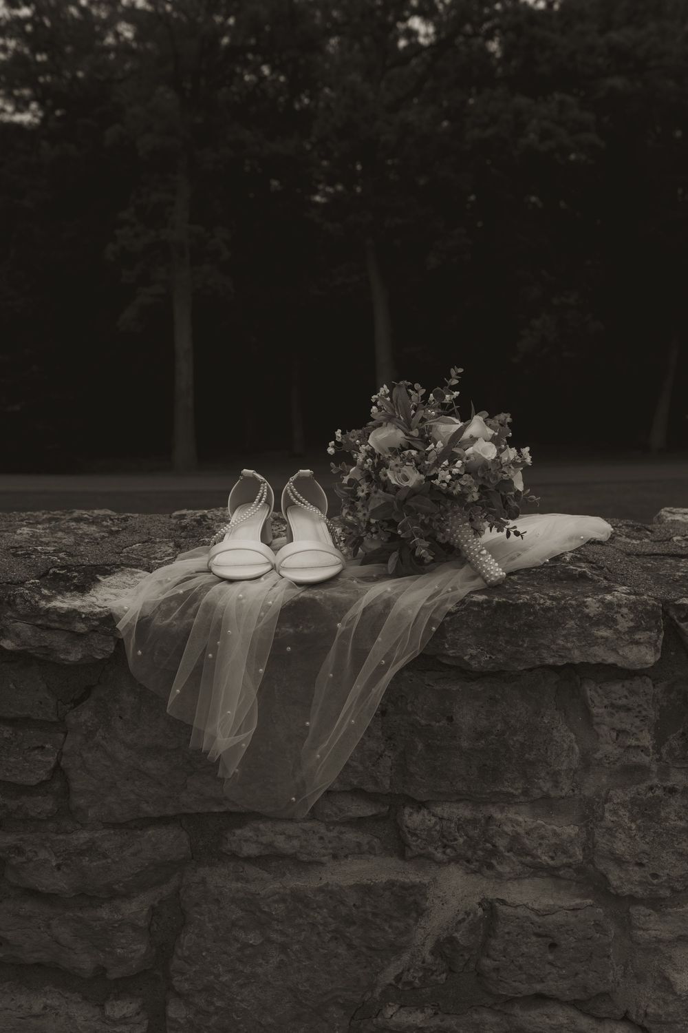 Black and white image of bridal accessories and bouquet arranged on stone wall outdoors.