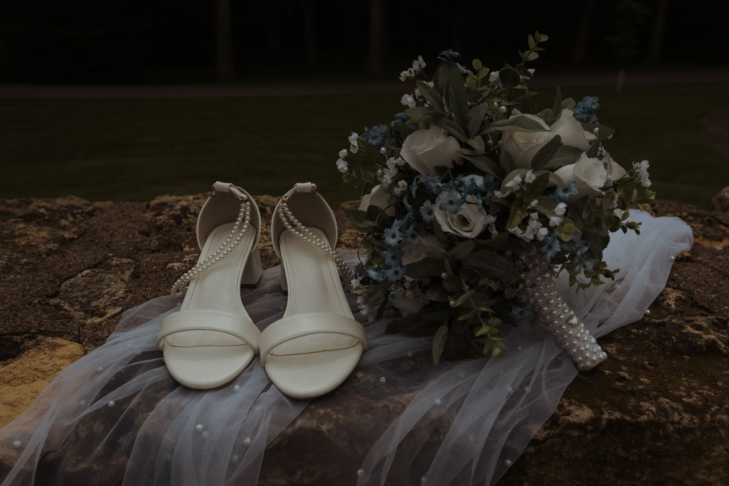 Wedding shoes and floral bouquet artistically arranged on stone wall with tulle veil.