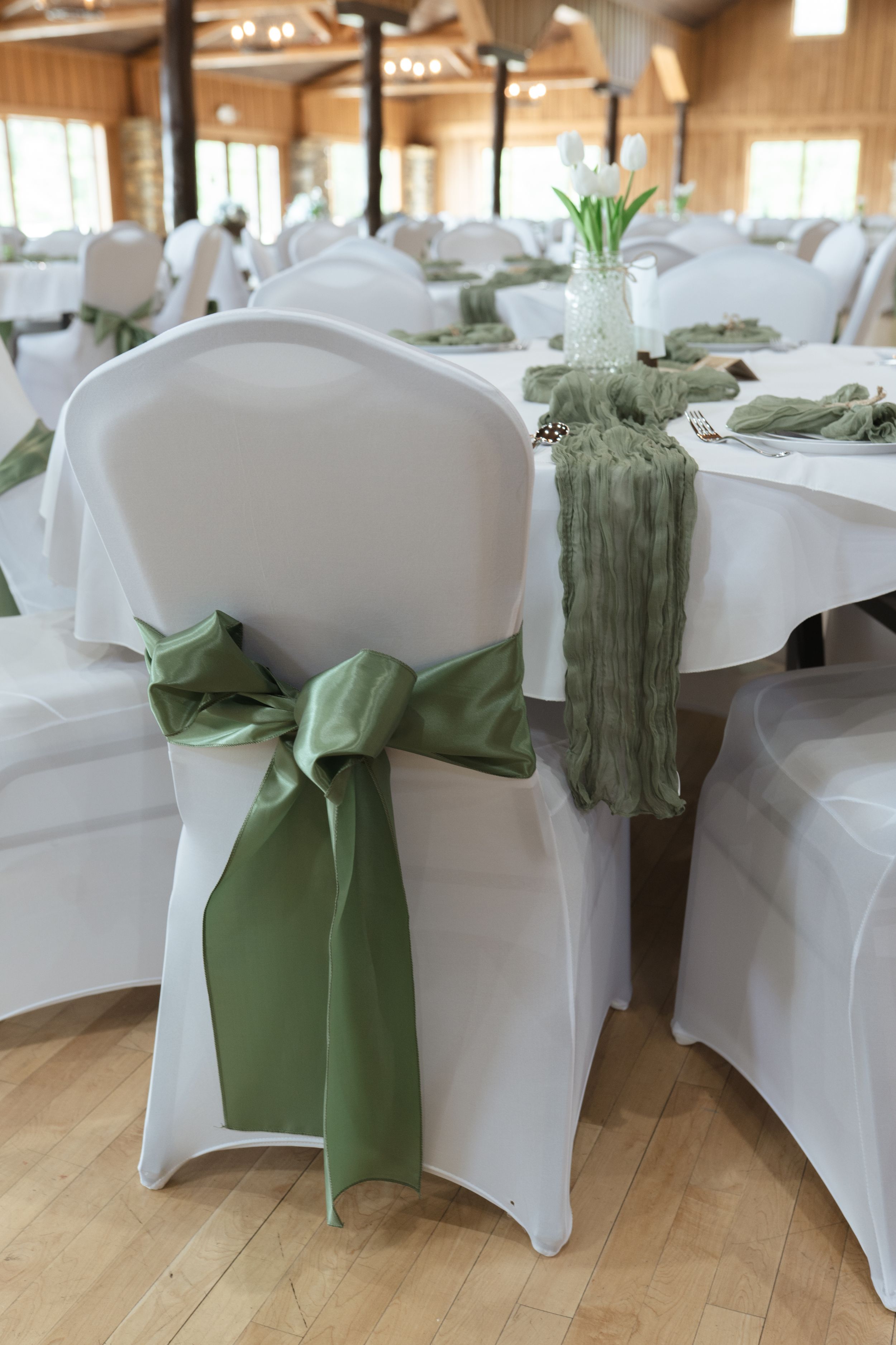 Reception venue with white chair covers and sage green sashes in a rustic wooden hall.