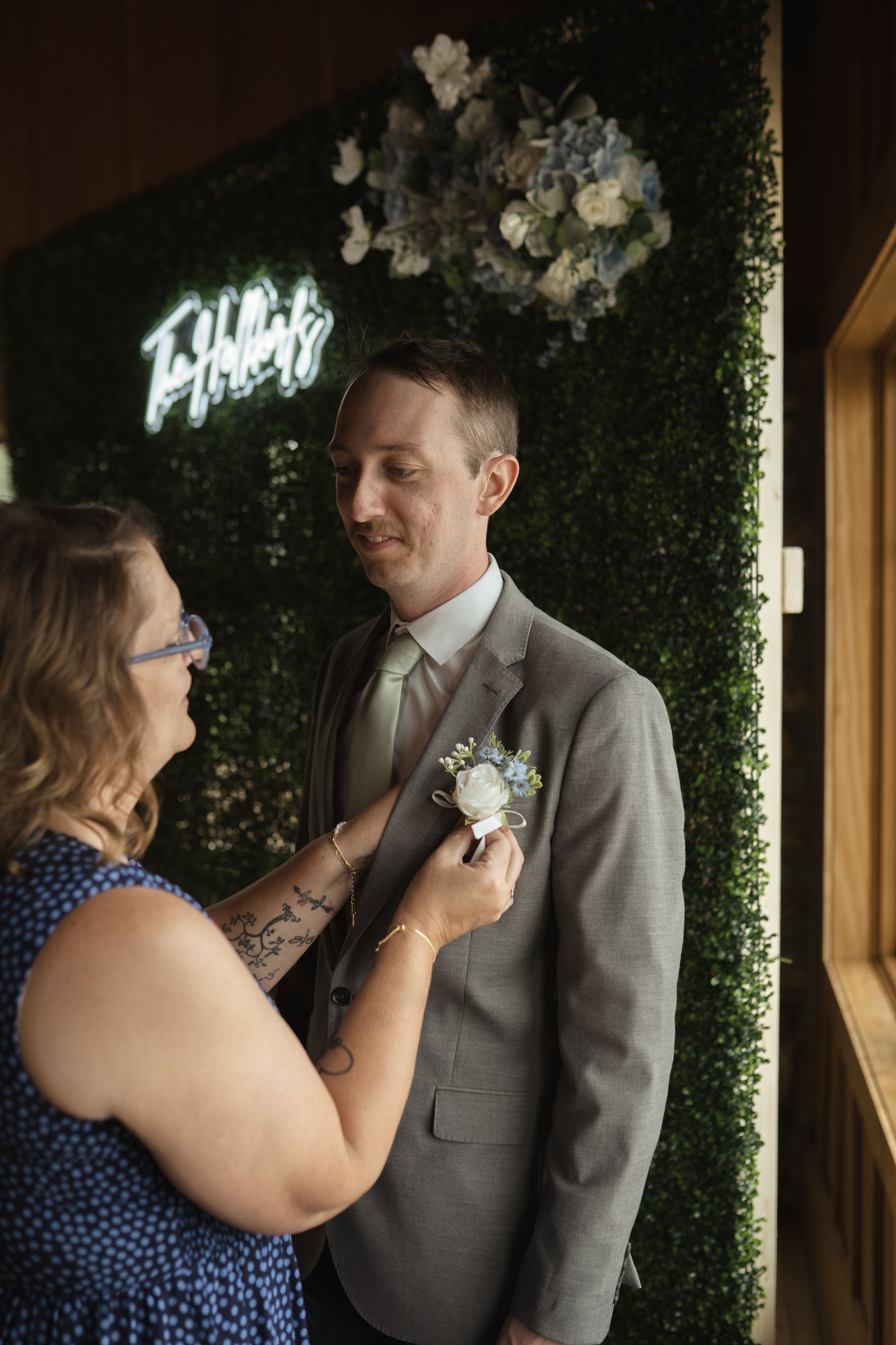 Wedding preparation moment in front of a green hedge wall with neon signage.
