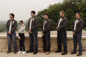 Groomsmen in dark suits and jeans pose casually against a stone wall outdoors.