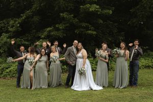 Wedding party members in sage green and gray attire sit casually on grass for group photo.